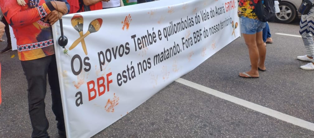 A group of indigenous people protest with a large banner on a street in a town: the banner reads: ‘The Tembé and Quilombola peoples of the Acará Valley are crying out. BBF is killing us. BBF out of our territory