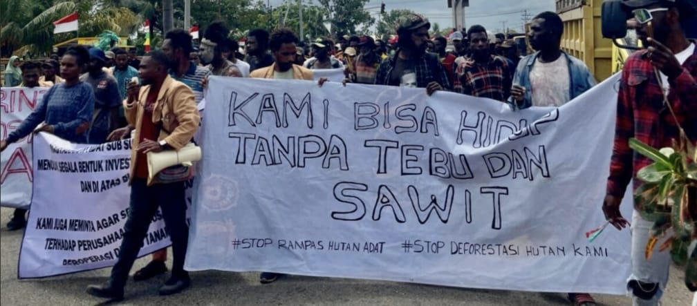 Crowd with banner: “We can live without sugar and palm oil. Stop the theft of Indigenous land. Stop the destruction of our forest”