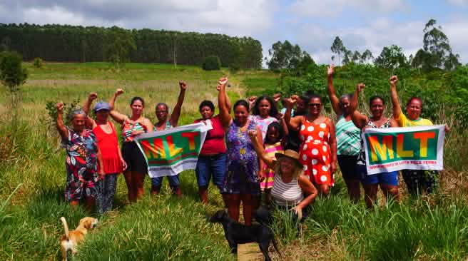A group of 15 women is protesting with their fists raised and two banners bearing the letters MLT