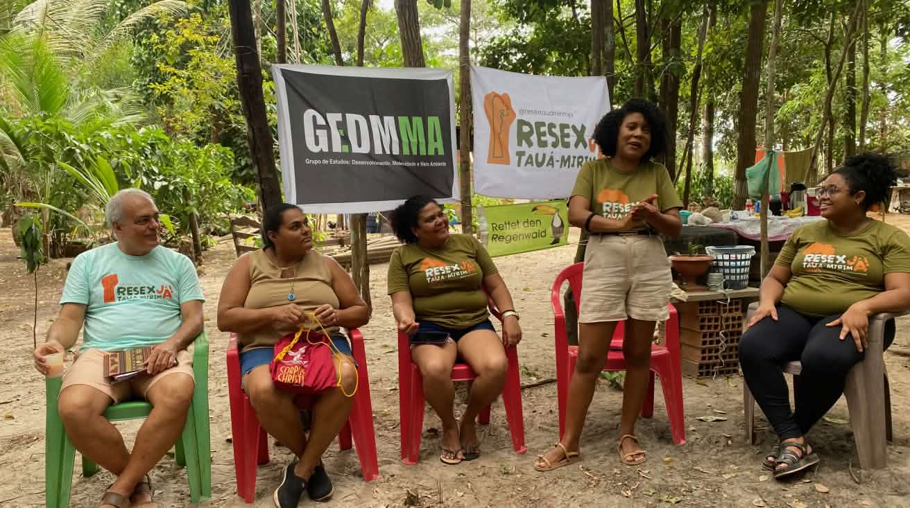 A woman speaks surrounded by four people sitting on chairs under trees and in front of three banners stretched across the tree trunks