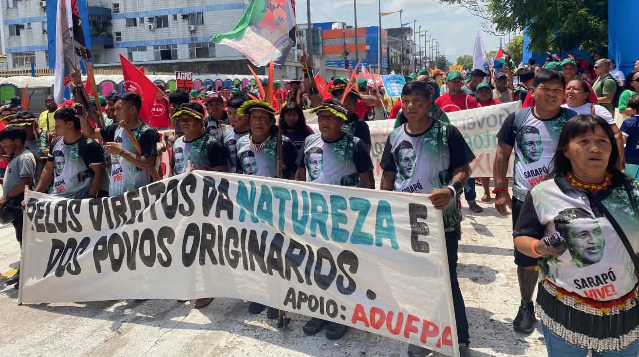 A group of around 15 Indigenous people with feather headdresses and a large banner