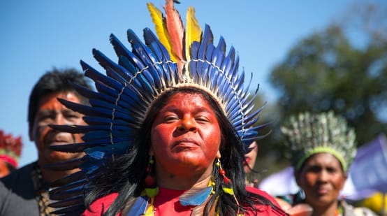 An indigenous woman adorned with a feathered crown in front of two other indigenous people