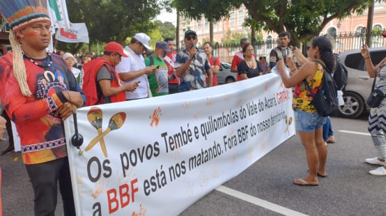 A group of indigenous people protest with a large banner on a street in a town: the banner reads: ‘The Tembé and Quilombola peoples of the Acará Valley are crying out. BBF is killing us. BBF out of our territory