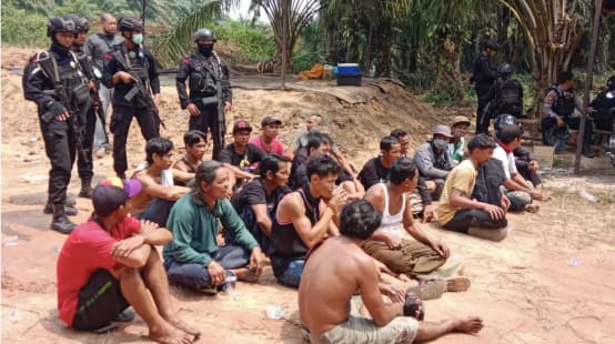 Detained Dayak people sit on the ground, armed police officers behind them