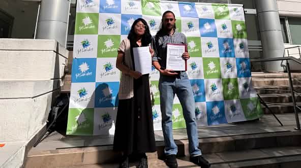 A woman and a man standing in front of a building entrance next to a banner reading “YASunidos,” holding documents in their hands