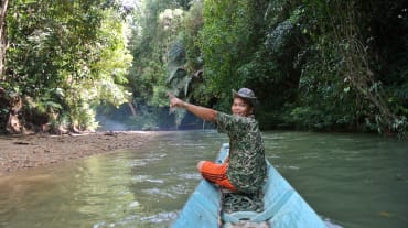 A smiling man at the helm of a small boat points toward the rainforest