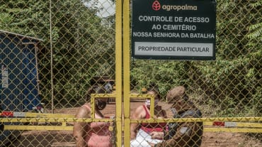 Two women behind a high barred gate are being checked by a guard. Inscription on the sign: Agropalma. Access control for Nossa Senhora da Batalha cemetery. Private property