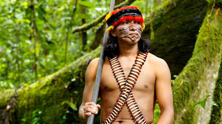 An indigenous man wearing feathers and a painted face standing in the forest with a blowpipe