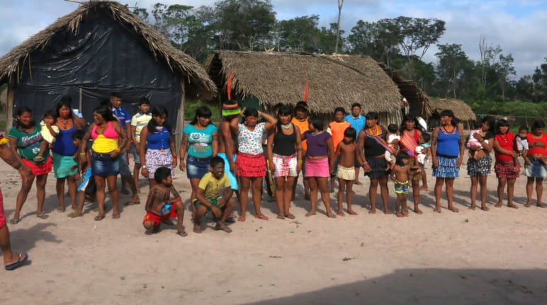 Indigenous Ka'apor standing in a double row in front of two huts in the forest