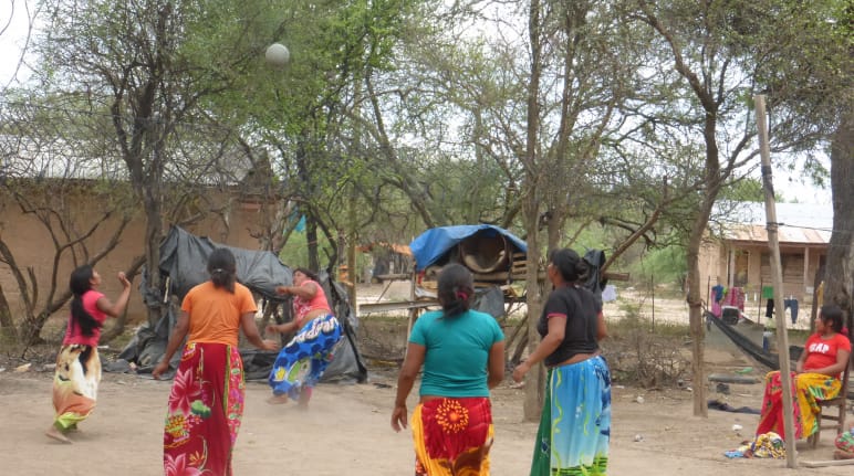Five indigenous women play a ball game between trees and wooden houses
