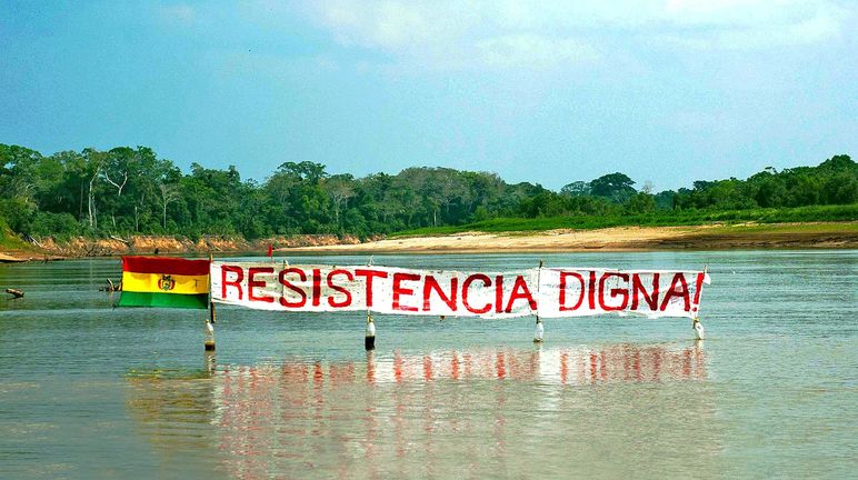 Inhabitants of the TIPNIS national park are protesting a planned road through the rainforest. Protest banner on a river in the Bolivian rainforest: the slogan “Resistance Means Dignity” stands next to the Bolivian flag.