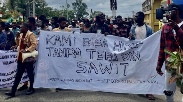 Crowd with banner: “We can live without sugar and palm oil. Stop the theft of Indigenous land. Stop the destruction of our forest”