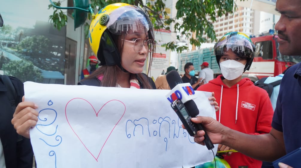 A young woman in a colorful helmet holds up a poster with a heart and speaks into a microphone