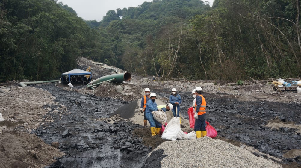 A group of oil workers work between two streams of black oil that have spilled down a boulder-covered mountainside in the rainforest. In the background, a section of pipe from the oil pipeline can be seen.