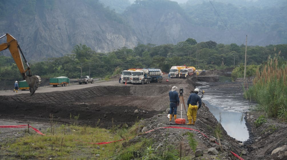Engineers measure the terrain between a pond filled with crude oil on the right and the shovel of an excavator visible on the left, which is digging another pit. In the background are tankers against the backdrop of mountains