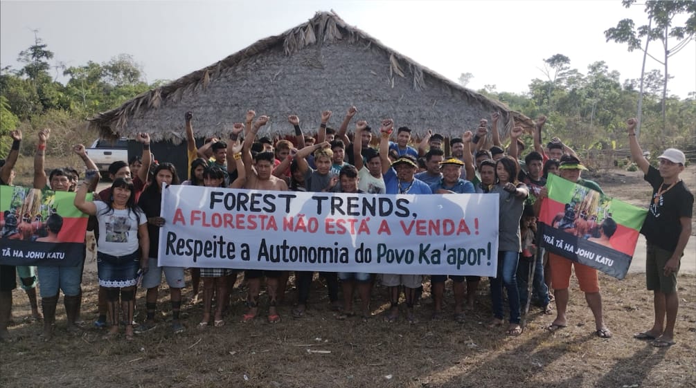 A group of Indigenous people with protest banners in front of a hut covered with palm fronds