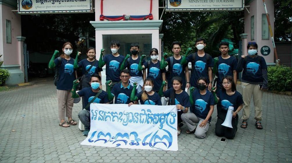 A group of 15 young people standing and kneeling in front of the Ministry of Tourism building, holding a banner to the camera. It reads “Koh Kong Island must become a marine national park”