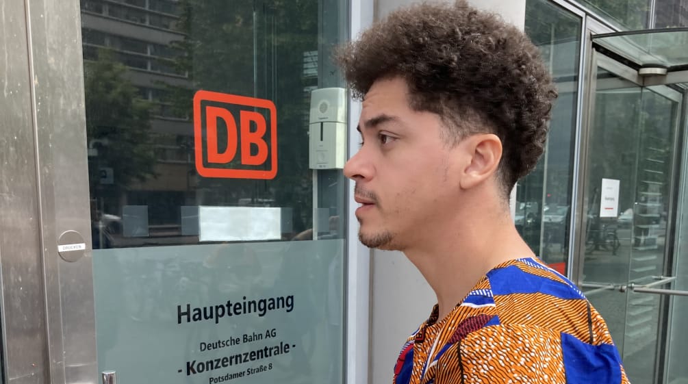 Mikaell from the Brazilian organization Justiça nos Trilhos in front of the DB headquarters in Berlin A young man stands with an envelope in front of a glass door with the inscription DB Main Entrance, Corporate Headquarters