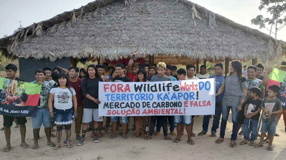 A group of Indigenous people with protest banners in front of a hut thatched with palm fronds