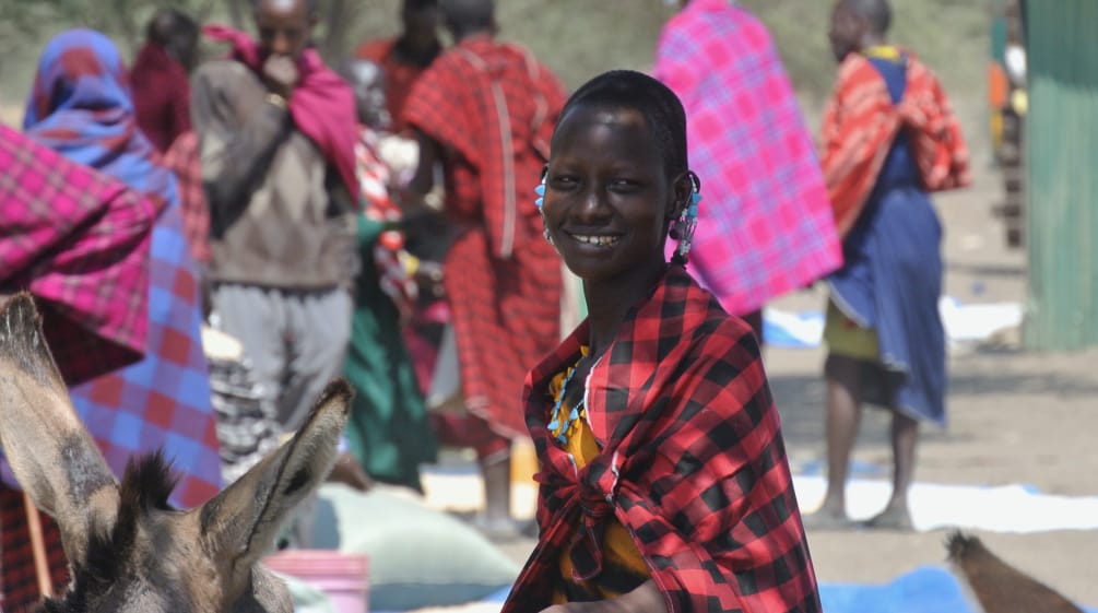 Maasai women and men in colorful cloths at a market, a woman looks at the camera