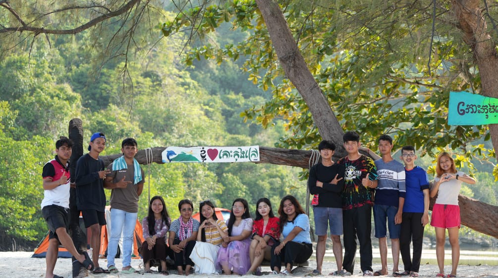 A group of young people sitting and standing in front of a tree on the beach