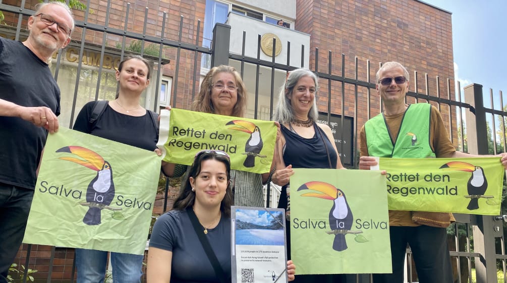 Six people standing in front of the Cambodian embassy compound's fence, holding banners with the organization’s toucan logo and the texts Rettet den Regenwald and Salva la Selva