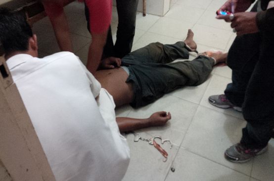 The corpse of a man lies on the floor. His head is not visible. Handcuffs are attached to one wrist and his feet are bound.