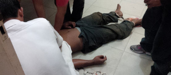 The corpse of a man lies on the floor. His head is not visible. Handcuffs are attached to one wrist and his feet are bound.