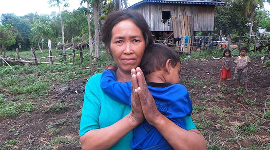 A woman holding a child, standing in front of their makeshift hut after being driven from their land
