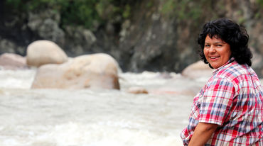Environmental activist Berta Cáceres sitting on a rock by a river, looking over her shoulder to the camera