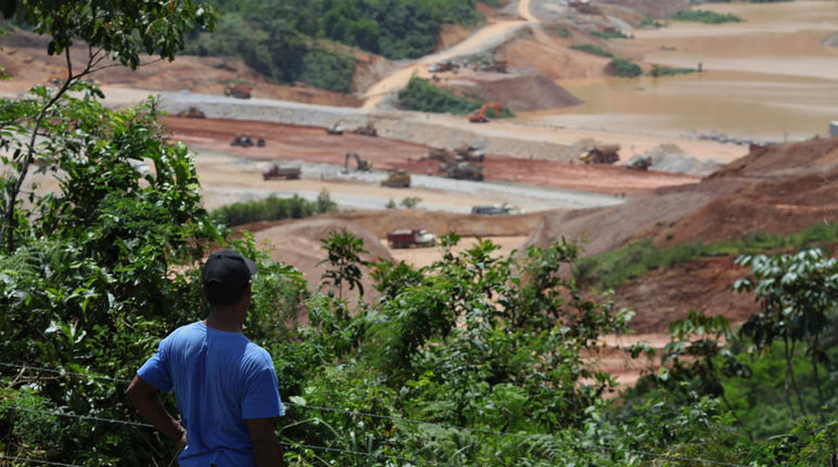 A farmer standing in the forest, looking at an open-pit mine