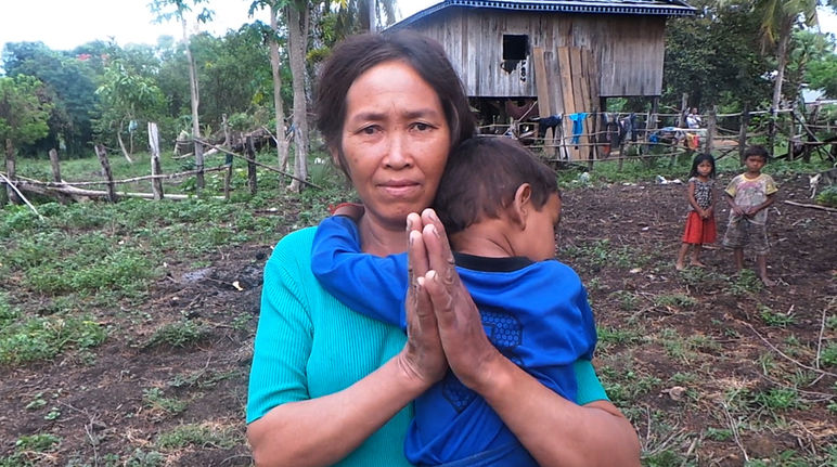 A woman holding a child, standing in front of their makeshift hut after being driven from their land